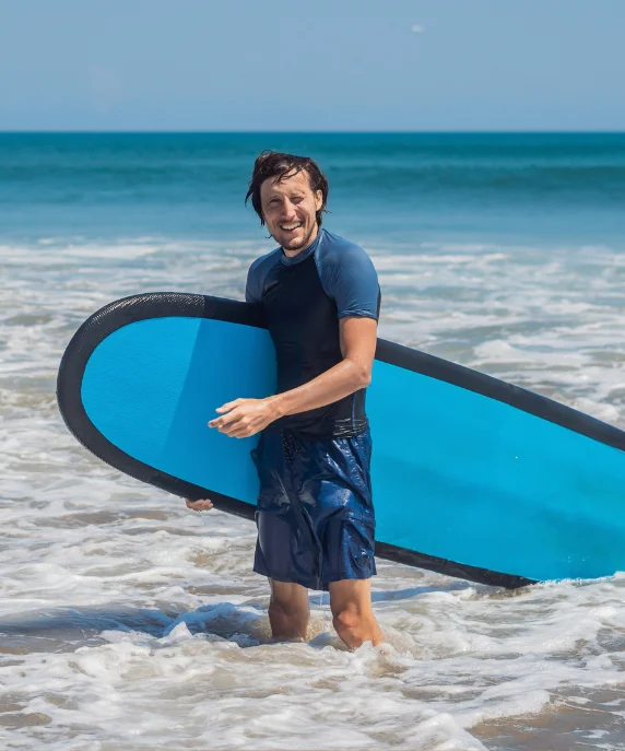 Homem sorrindo na beira do mar carregando uma prancha de iniciante azul